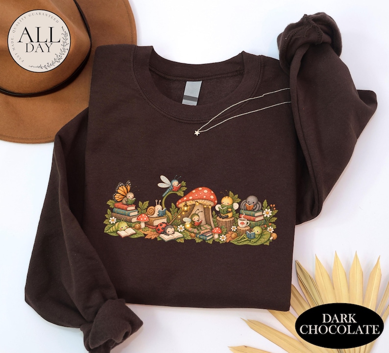 K&ouml;nnte beinhalten: Ein dunkelbraunes Sweatshirt mit Rundhalsausschnitt und einem farbenfrohen Grafikdruck, der verschiedene Insekten und kleine Lebewesen zeigt, die zwischen Pilzen, Bl&auml;ttern und Blumen B&uuml;cher lesen. Das Design umfasst einen Schmetterling, eine Schnecke, einen Marienk&auml;fer und eine Biene.