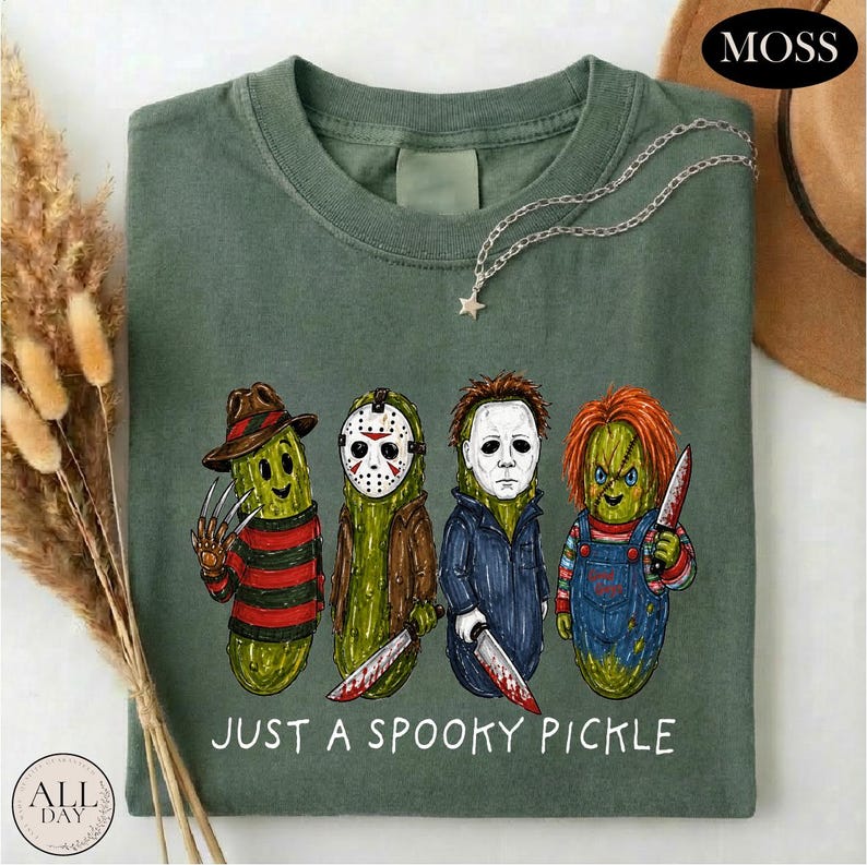 May include: A sage green t-shirt featuring a graphic of four anthropomorphic pickles dressed as horror movie characters. Below the characters, the text reads "JUST A SPOOKY PICKLE" in white capital letters. The shirt has a crew neckline.