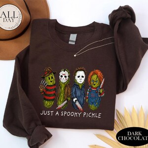 May include: A dark brown crewneck sweatshirt featuring a graphic of four green pickles dressed as horror movie characters. Below the characters, the text reads "JUST A SPOOKY PICKLE" in white letters. The sweatshirt has long sleeves and a ribbed collar.