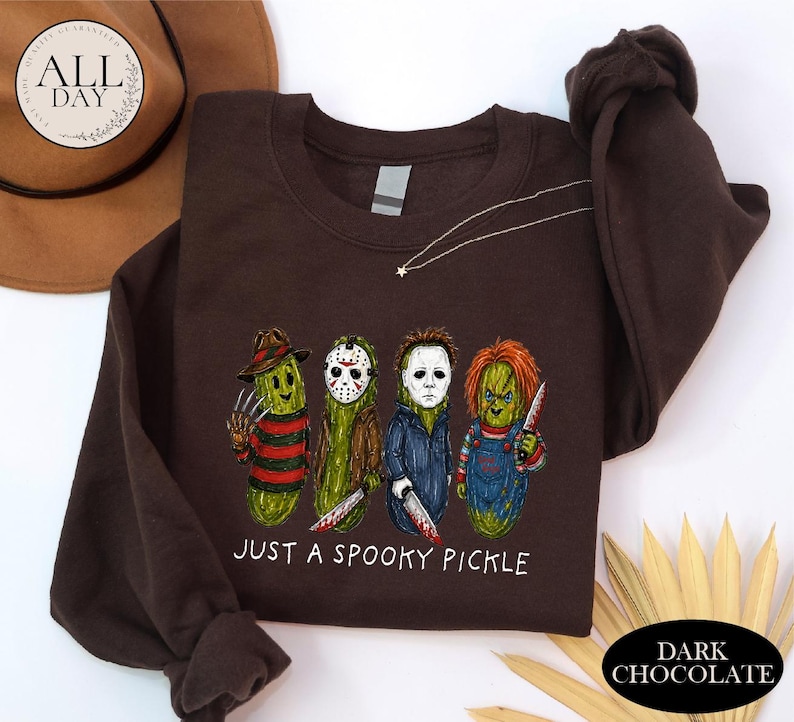 May include: A dark brown crewneck sweatshirt featuring a graphic of four green pickles dressed as horror movie characters. Below the characters, the text reads "JUST A SPOOKY PICKLE" in white letters. The sweatshirt has long sleeves and a ribbed collar.