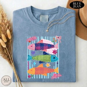 May include: A blue crew neck t-shirt featuring a graphic of three stacked fish in plaid and checkered patterns. The design includes small decorative elements like stars, a heart, and a bow. The shirt has a faded, washed-out appearance.