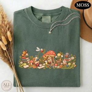 K&ouml;nnte beinhalten: Ein waldgr&uuml;nes T-Shirt mit einer grafischen Illustration verschiedener kleiner Insekten und Kreaturen, die unter einem gro&szlig;en roten Pilz B&uuml;cher lesen. Das Design zeigt einen Schmetterling, eine Schnecke, einen Marienk&auml;fer, eine Biene und eine Spinne.