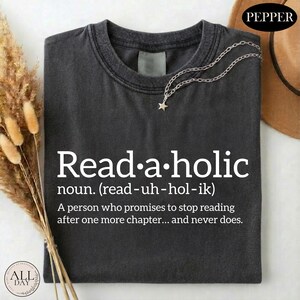 May include: A charcoal gray t-shirt featuring white text that reads Read-a-holic noun. (read-uh-hol-ik) A person who promises to stop reading after one more chapter... and never does. A silver-toned chain necklace with a small star charm rests on the collar.