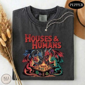May include: Dark gray t-shirt featuring a fantasy scene with dragons and the text "HOUSES & HUMANS" in red. The graphic depicts four dragons around a table with miniature figures and a campfire. A silver necklace with a star pendant is draped on the shirt.