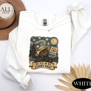 May include: A white long-sleeve sweatshirt featuring a graphic of a black cat wearing a hat, set against a swirling starry night background. The cat holds a sign that says NO ICE and a banner below reads NO ICE IN MY STARRY NIGHT.