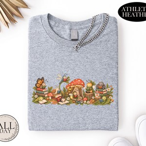 K&ouml;nnte beinhalten: Ein athletisch graues T-Shirt mit einer grafischen Illustration verschiedener Insekten, die unter einem gro&szlig;en roten Pilz B&uuml;cher lesen. Das Design zeigt einen Schmetterling, eine Schnecke, einen Marienk&auml;fer, eine Biene und eine Spinne zwischen B&uuml;chern.