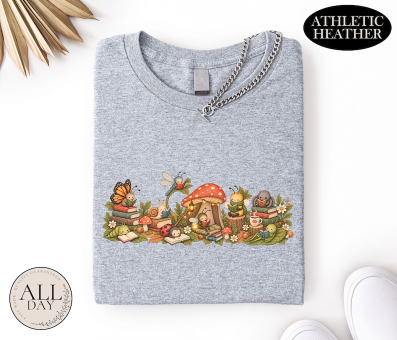 K&ouml;nnte beinhalten: Ein athletisch graues T-Shirt mit einer grafischen Illustration verschiedener Insekten, die unter einem gro&szlig;en roten Pilz B&uuml;cher lesen. Das Design zeigt einen Schmetterling, eine Schnecke, einen Marienk&auml;fer, eine Biene und eine Spinne zwischen B&uuml;chern.