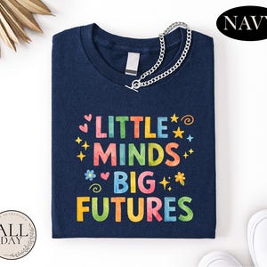 A navy blue t-shirt with the text "LITTLE MINDS BIG FUTURES" printed on it, along with colorful stars and hearts. The t-shirt is folded and placed on a white surface, with a navy blue "NAVY" logo visible in the top right corner.