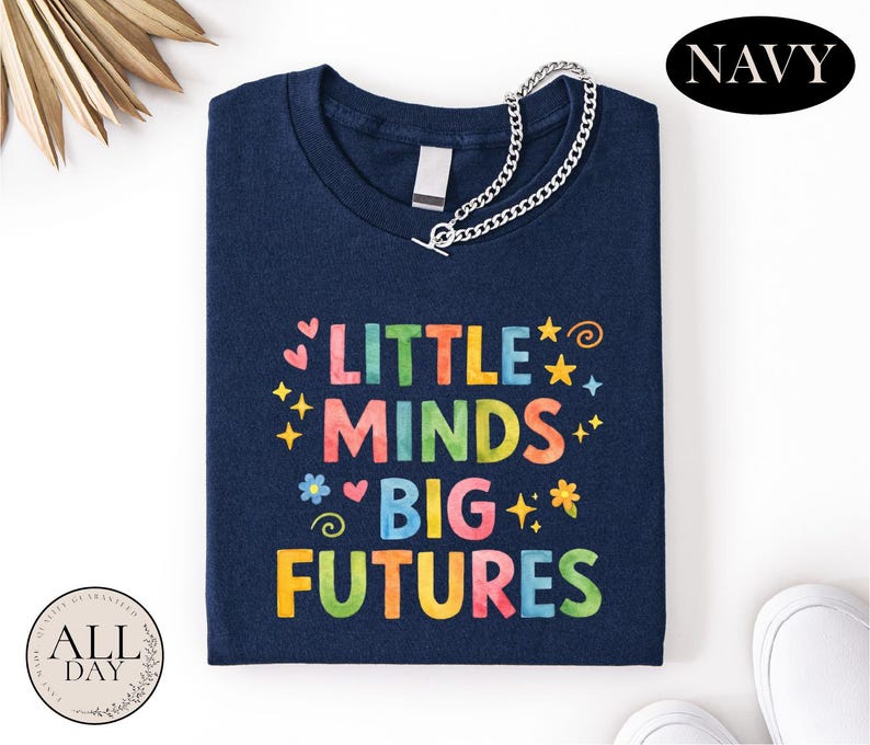 A navy blue t-shirt with the text "LITTLE MINDS BIG FUTURES" printed on it, along with colorful stars and hearts. The t-shirt is folded and placed on a white surface, with a navy blue "NAVY" logo visible in the top right corner.