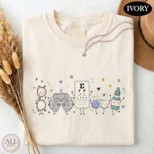 May include: An ivory t-shirt featuring a graphic print of five anthropomorphic eye care items: a pair of glasses, a phoropter, an eye chart with the letters E, F, P, a contact lens case, and an eye drop bottle, all surrounded by small colorful stars.