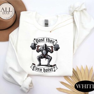 a white long-sleeved shirt with a graphic design depicting a knight lifting a barbell, accompanied by the text "Post thou Even hoist?"