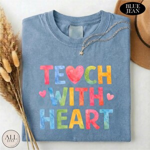 A blue t-shirt with the text "TEACH WITH HEART" printed on it, along with colorful heart shapes. The t-shirt is folded and placed on a white surface, with some dried flowers and a brown hat nearby.