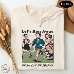 May include: An ivory t-shirt featuring a graphic print of two children and a dog running outdoors. The text above the illustration reads Let's Run Away and the text below reads FROM OUR PROBLEMS. The shirt has a crew neckline and short sleeves.