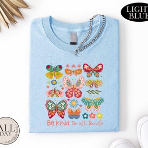 A light blue t-shirt with a colorful butterfly design and the text "Be Kind to all kinds" printed on it.