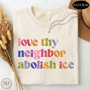 May include: Cream-colored t-shirt with the words "love thy neighbor abolish ice" in colorful, retro-style lettering. A silver necklace with a star pendant rests on the shirt. The shirt is folded and displayed on a flat surface.