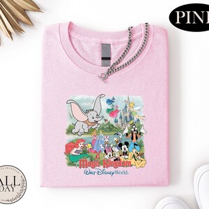 A pink t-shirt with a Disney-themed graphic, including characters from the Magic Kingdom, is displayed on a white background.
