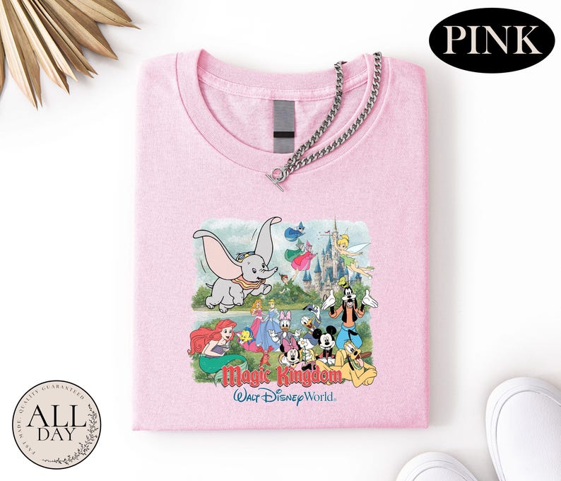 A pink t-shirt with a Disney-themed graphic, including characters from the Magic Kingdom, is displayed on a white background.