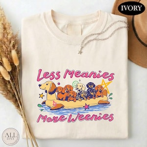 May include: An ivory t-shirt featuring a graphic of multiple dachshund dogs riding a yellow inflatable pool float. The text "Less Meanies More Weenies" is printed in pink above and below the illustration, accompanied by small star and smiley face icons.