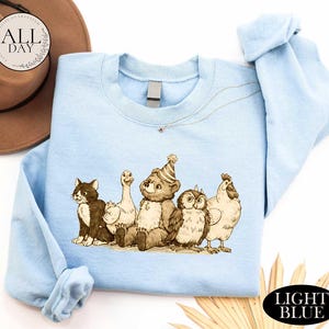 A light blue t-shirt with a cartoon illustration of a bear, cat, duck, chicken, and owl, along with a brown hat and straw hat, all set against a white background.