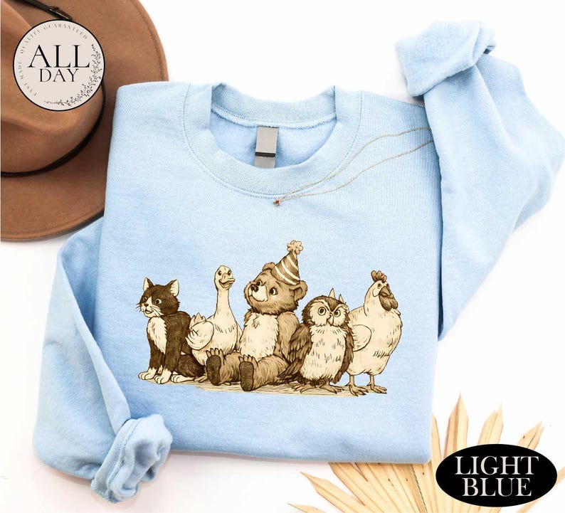 A light blue t-shirt with a cartoon illustration of a bear, cat, duck, chicken, and owl, along with a brown hat and straw hat, all set against a white background.