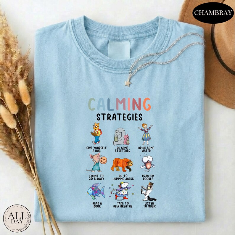 A light blue t-shirt with a colorful graphic design depicting various calming strategies, such as giving oneself a hug, drinking water, and reading a book.