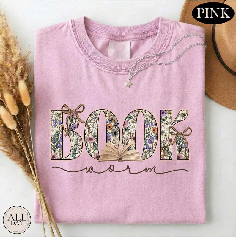 A pink t-shirt with the word "BOOK" printed on it, surrounded by floral designs. The t-shirt is folded and placed on a white surface, with some dried flowers and a hat visible in the background.