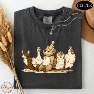 a black t-shirt with a graphic design depicting a group of animals, including a cat, duck, chicken, and owl, sitting together.