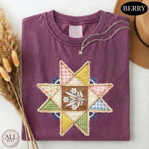 May include: A berry-colored t-shirt featuring a central star-shaped quilt pattern. The star is composed of various fabric patches in gingham, floral, and solid prints. A delicate silver-toned chain necklace with a small star charm rests on the collar.