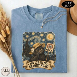 May include: A blue jean colored t-shirt featuring a graphic of a black cat wearing a straw hat. The cat holds a sign that says NO ICE with a crossed-out ice cube icon. Below the cat, a banner reads NO ICE IN MY STARRY NIGHT in dark text.