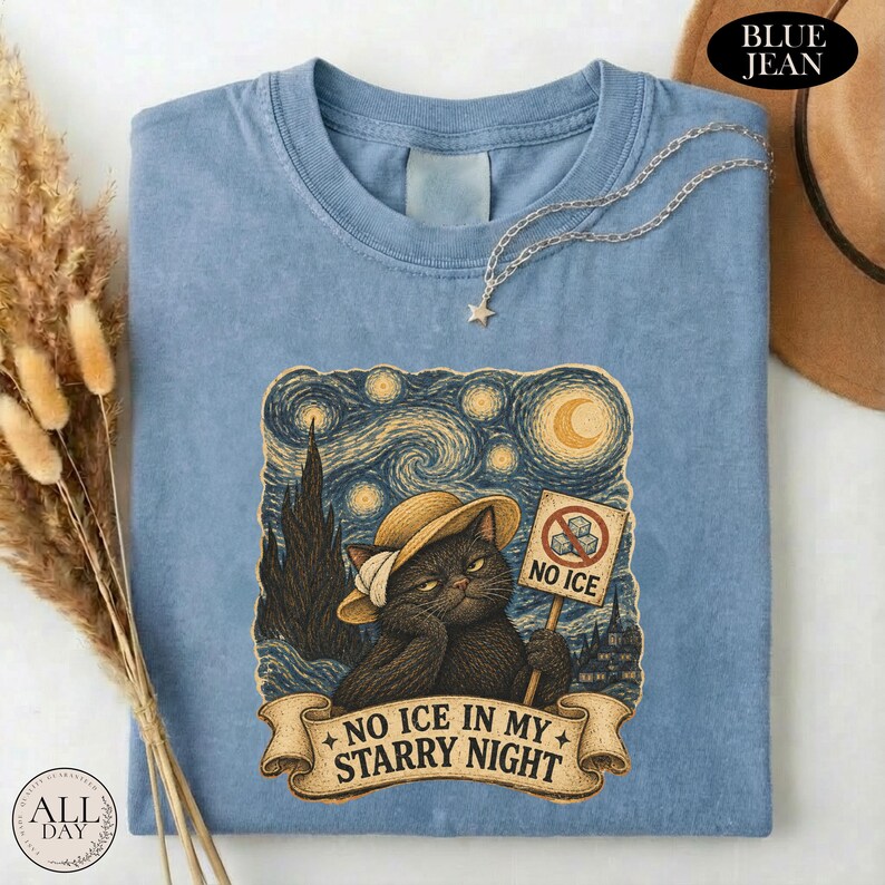 May include: A blue jean colored t-shirt featuring a graphic of a black cat wearing a straw hat. The cat holds a sign that says NO ICE with a crossed-out ice cube icon. Below the cat, a banner reads NO ICE IN MY STARRY NIGHT in dark text.