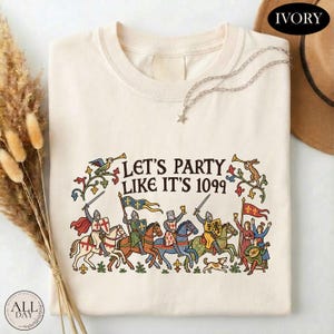 May include: An ivory t-shirt featuring a colorful medieval-themed graphic with knights on horseback, musicians, and the text "LET'S PARTY LIKE IT'S 1099". The design includes detailed illustrations of figures in armor, flags, and floral motifs in red, blue, and gold.