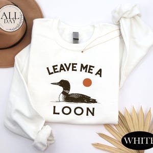 a white long-sleeved t-shirt with a graphic design of a duck and the text "LEAVE ME A LON" printed on it. The t-shirt is laid out on a white surface, along with a hat and some straws.