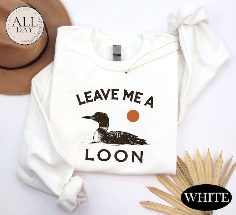 a white long-sleeved t-shirt with a graphic design of a duck and the text "LEAVE ME A LON" printed on it. The t-shirt is laid out on a white surface, along with a hat and some straws.