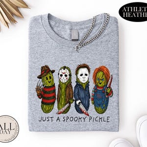 May include: An athletic heather gray t-shirt featuring a graphic of four green pickles dressed as horror movie characters. Below the characters, the text reads "JUST A SPOOKY PICKLE". The shirt has a crew neckline and a silver chain necklace attached.