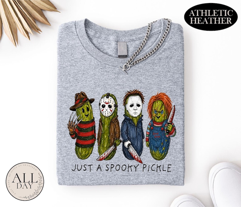 May include: An athletic heather gray t-shirt featuring a graphic of four green pickles dressed as horror movie characters. Below the characters, the text reads "JUST A SPOOKY PICKLE". The shirt has a crew neckline and a silver chain necklace attached.