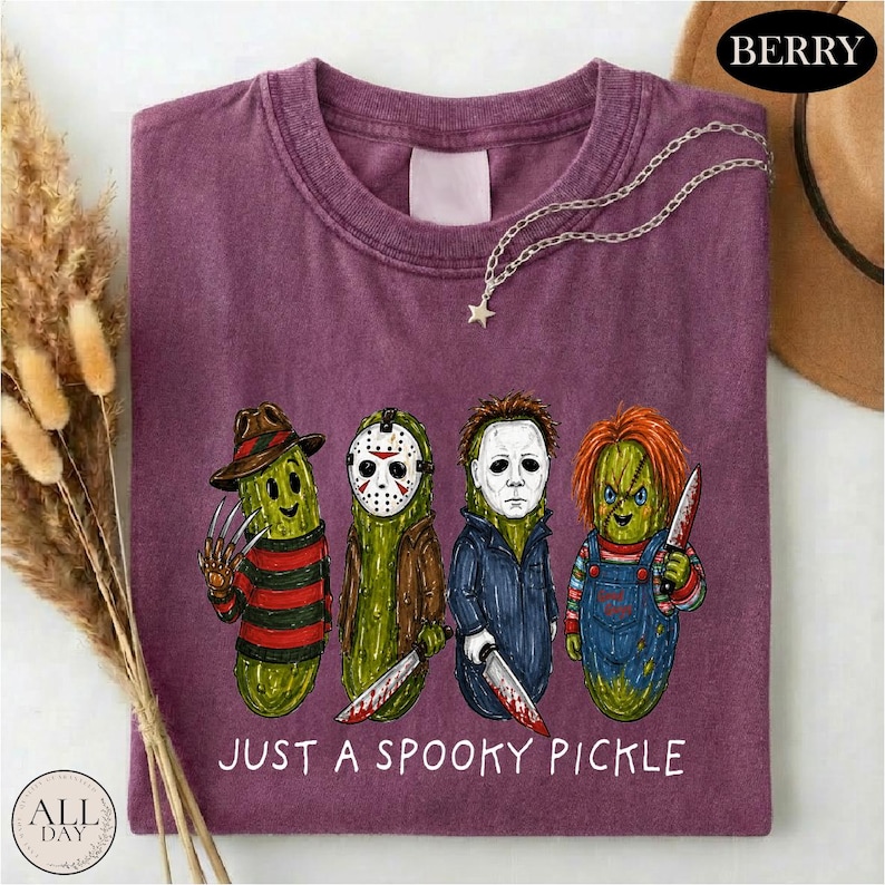 May include: A berry-colored t-shirt featuring a graphic of four green pickles dressed as horror movie characters. The text below the characters reads JUST A SPOOKY PICKLE. The shirt has a crew neckline and a short-sleeved design.