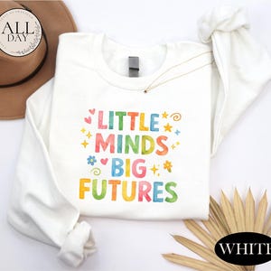 a white long-sleeved shirt with a colorful text design that says "LITTLE MINDS BIG FUTURES" printed on it. The shirt is laid out on a white background, and there is a brown hat and some straws in the background.