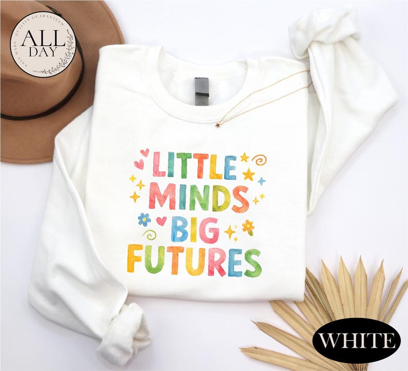 a white long-sleeved shirt with a colorful text design that says "LITTLE MINDS BIG FUTURES" printed on it. The shirt is laid out on a white background, and there is a brown hat and some straws in the background.