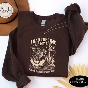 a brown sweatshirt with a graphic design depicting a dragon and castle, along with the text "I HAD THE TIME OF MY LIFE RIDING DRAGONTS WITH YOU". The sweatshirt is placed on a white surface, with a hat and some leaves visible in the background.