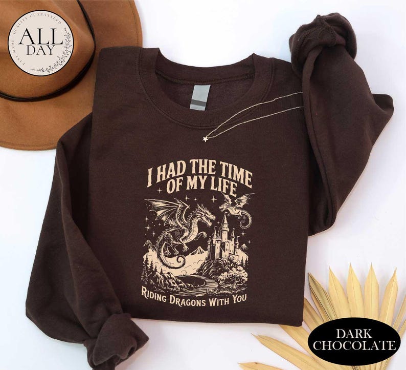 a brown sweatshirt with a graphic design depicting a dragon and castle, along with the text "I HAD THE TIME OF MY LIFE RIDING DRAGONTS WITH YOU". The sweatshirt is placed on a white surface, with a hat and some leaves visible in the background.