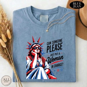 May include: A blue crew neck t-shirt featuring a graphic of the Statue of Liberty wearing star-spangled sunglasses. The text reads: CAN SOMEONE PLEASE JUST PUT A WOMAN IN CHARGE? The design includes red and blue accents and small star icons.