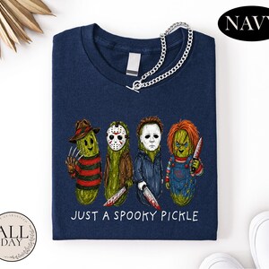 May include: A navy blue t-shirt featuring a graphic of four pickles dressed as horror movie characters. Below the pickles, the text reads "JUST A SPOOKY PICKLE" in white. The shirt has a silver chain attached to the neckline and a blank tag.