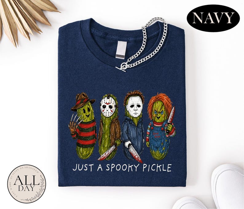 May include: A navy blue t-shirt featuring a graphic of four pickles dressed as horror movie characters. Below the pickles, the text reads "JUST A SPOOKY PICKLE" in white. The shirt has a silver chain attached to the neckline and a blank tag.