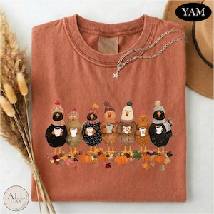 May include: A rust-colored t-shirt featuring a graphic of seven chickens wearing knit hats and scarves, each holding a white mug. The chickens stand above a pile of autumn leaves and small orange pumpkins. A silver star necklace rests on the shirt.