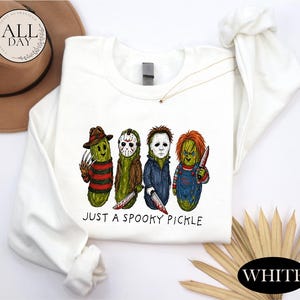 May include: A white long-sleeve sweatshirt featuring a graphic of four green pickles dressed as horror movie characters. Below the characters, the text reads "JUST A SPOOKY PICKLE" in black lettering. The design includes iconic masks, hats, and weapons.