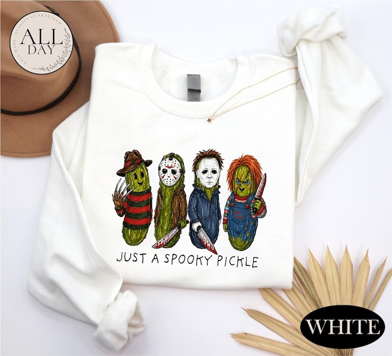 May include: A white long-sleeve sweatshirt featuring a graphic of four green pickles dressed as horror movie characters. Below the characters, the text reads "JUST A SPOOKY PICKLE" in black lettering. The design includes iconic masks, hats, and weapons.