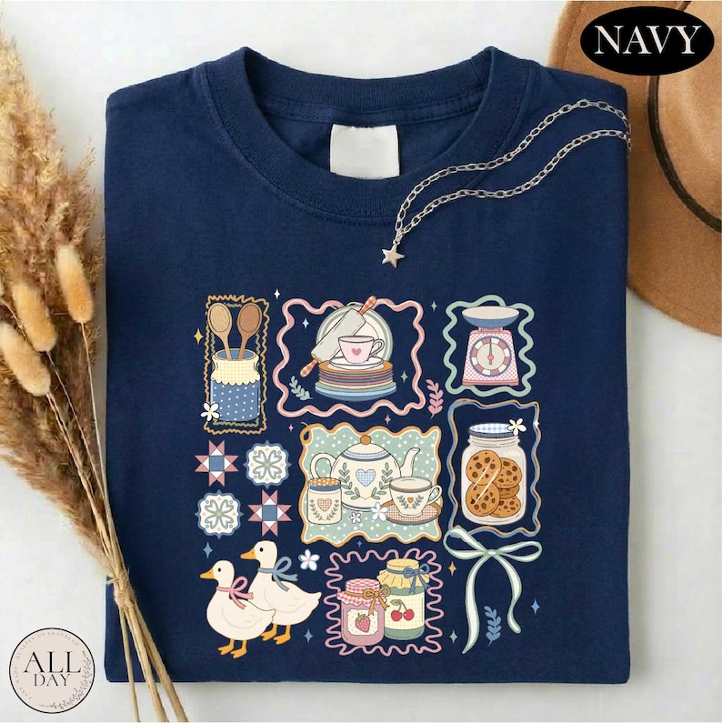 a navy blue t-shirt with a colorful and whimsical design, including illustrations of various kitchen items and objects such as a teapot, spoons, a cake, and a clock. The t-shirt is displayed on a white surface, and there is a brown hat placed next to it.
