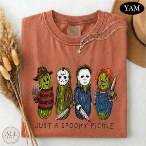 May include: A rust-colored t-shirt featuring a graphic of four green pickles dressed as horror movie characters. Below the illustration, the text reads JUST A SPOOKY PICKLE. The shirt has a crew neckline and short sleeves, presented in a flat lay.