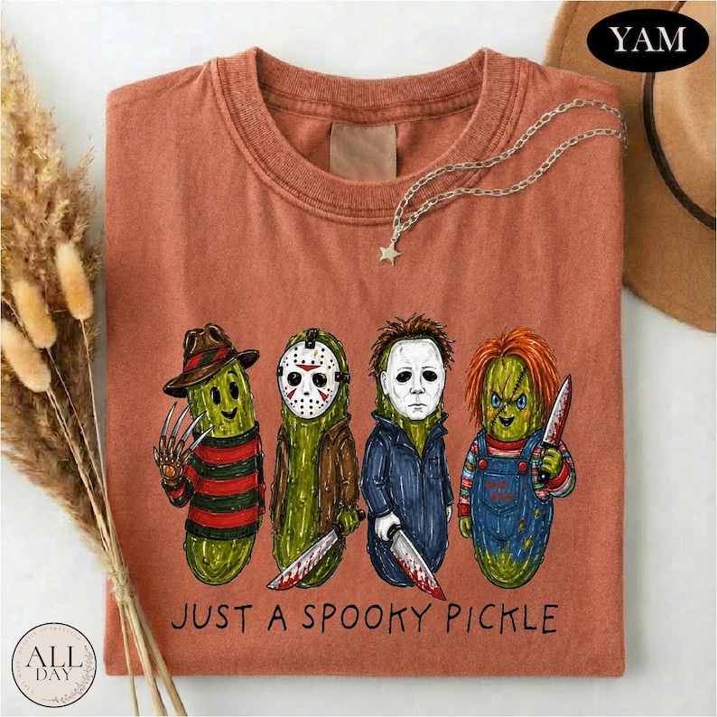 May include: A rust-colored t-shirt featuring a graphic of four green pickles dressed as horror movie characters. Below the illustration, the text reads JUST A SPOOKY PICKLE. The shirt has a crew neckline and short sleeves, presented in a flat lay.