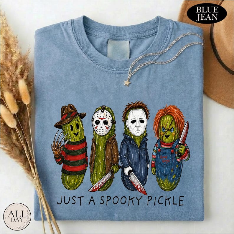 May include: A light blue t-shirt featuring a graphic of four pickles dressed as horror movie characters. The text below the characters reads JUST A SPOOKY PICKLE. The shirt has a crew neckline and a short-sleeved design with a casual, relaxed fit.